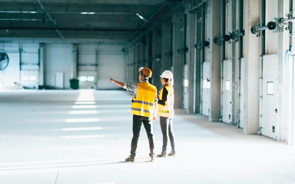 man and woman wearing yellow construction vests standing in large empty warehouse building